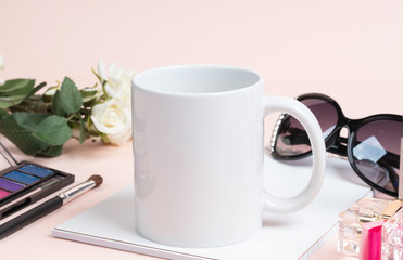 Obraz na płótnie Canvas White plain coffee mug on notebook with female fashion makeup accessory. Surrounded with perfume, sunglasses and white floor.