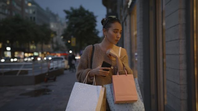 A young shopaholic woman with shopping bags at sunset and looking through a shop window