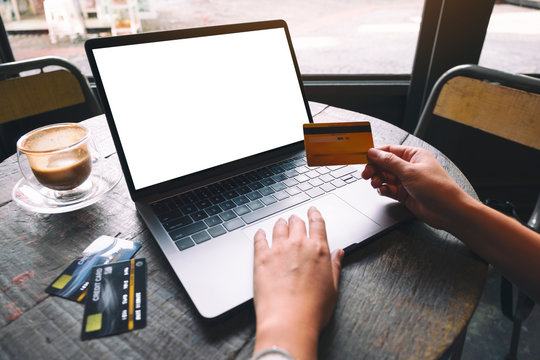 Mockup image of a hand holding credit card while using and typing on laptop with blank white screen and coffee cup on wooden table