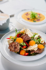 Business lunch - fresh vegetable salad with grilled meat and pumpkin cream soup in white plates on a white table in a restaurant. Close-up. Space