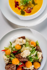 Business lunch - coffee, pumpkin cream soup and fresh vegetable salad with grilled meat in white plates on a white table in a restaurant. Close-up. Space. Flat lay. Top view