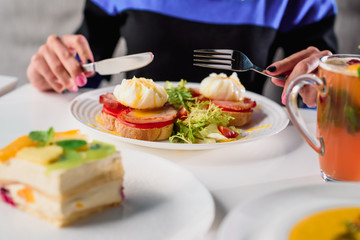 Woman eating toast with ham, tomato, poached egg and fresh salad in a restaurant. Healthy breakfast. Close-up. Space