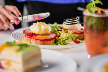 Woman eating toast with ham, tomato, poached egg and fresh salad in a restaurant. Healthy breakfast. Close-up. Space