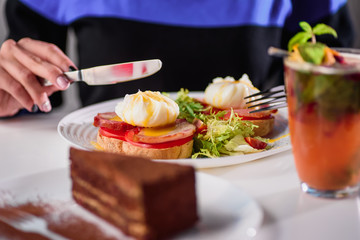 Woman eating toast with ham, tomato, poached egg and fresh salad in a restaurant. Healthy breakfast. Close-up. Space