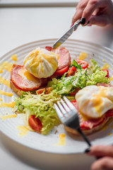 Woman eating toast with ham, tomato, poached egg and fresh salad in a restaurant. Healthy breakfast. Close-up. Space