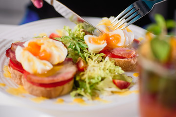 Woman eating toast with ham, tomato, poached egg and fresh salad in a restaurant. Healthy breakfast. Close-up. Space