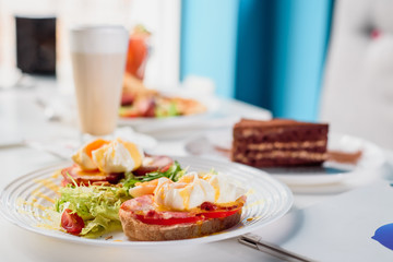 Toasts with ham, tomato and poached egg, served with fresh salad on white plate. Healthy breakfast on the table in the restaurant. Close-up. Space