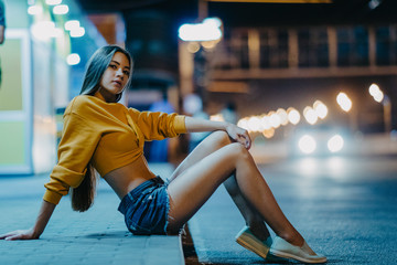 A woman sits on sidewalk against background of night city.