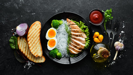 Rice noodles with chicken fillet, egg and broccoli in a black plate on a black background. Top view. Free space for your text.
