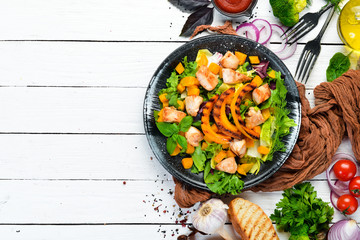 Pumpkin salad with chicken meat and vegetables in a black plate on a white wooden background. Top view. Free space for your text.