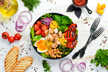 Buddha bowl: Chicken, chickpeas, chickpeas, broccoli, tomatoes, egg, paprika, onion in a black plate on a white wooden background. Top view. Free space for your text.