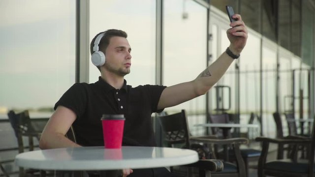 Young man with headphones on head doing selfie uses phone sitting in cafe