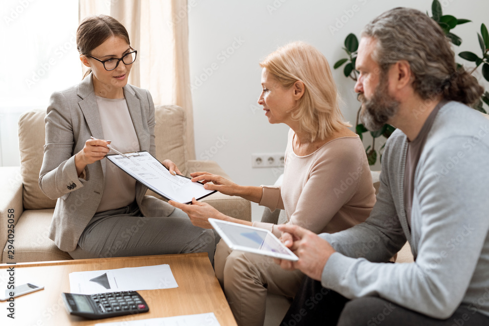 Poster young real estate agent pointing at document while consulting mature clients