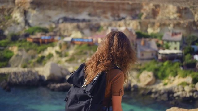 A young woman tourist with a backpack walks on a cliff on the background of a fishing village and considers nature.