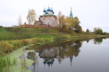 Obraz premium Old blue domes church and bellfray among colorful autumn trees reflecting in calm river water in cloudy day in Dunilovo village