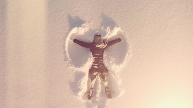 Aerial top view of young happy woman lying in snow and making snow angel by hands and legs in sunlight, toned
