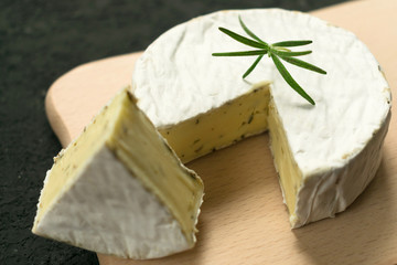  Camembert cheese with greens decorated with a sprig of rosemary on a wooden board on a black background.