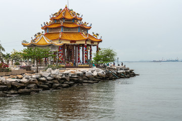 Obraz premium Si Racha, Thailand - March 16, 2019: Red roofed Guan Yin circular shrine on edge of Ko Loi island surrounded by flat see of Bay of Thailand and light blue sky. Some green foliage.