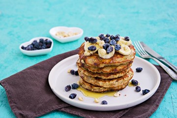 Banana oat pancakes with hazelnuts, sliced banana, honey and honeysuckle on a white plate on a turquoise concrete background. American food. Baking with oatmeal.