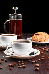 Two cups of coffee in a saucer and coffee beans on a dark wooden table. Croissant and coffeemaker in the background.