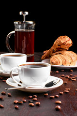 Two cups of coffee in a saucer and coffee beans on a dark wooden table. Croissant and coffeemaker in the background.
