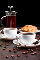 Two cups of coffee in a saucer and coffee beans on a dark wooden table. Croissant and coffeemaker in the background.