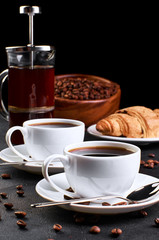 Two cups of coffee on a saucer and coffee beans on a dark background. In the background there is a croissant, a coffee pot and a wooden dish with coffee beans.
