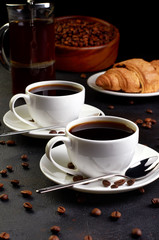 Two cups of coffee on a saucer and coffee beans on a dark background. In the background there is a croissant, a coffee pot and a wooden dish with coffee beans.