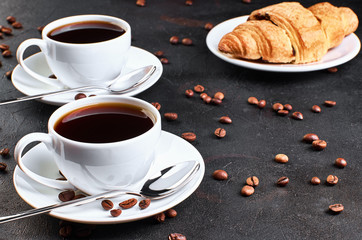 Two cups of coffee on a saucer and coffee beans on a dark background. Croissant in the background.