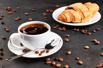 A Cup of coffee on a saucer and coffee beans on a dark background. Croissant in the background. A drop of coffee splashed in the Cup.