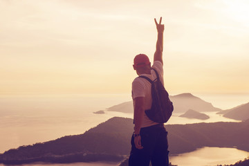 Fototapeta premium Young man standing on a hilltop with his hand raised up depicting a symbol of peace or victory, against a seascape at sunset, rear view, toned. Concept of freedom, happiness, success, victory