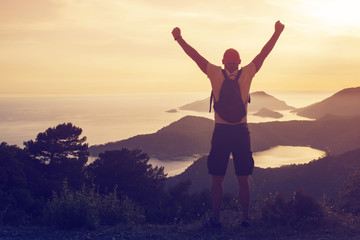 Young man with a backpack standing on top of a hill with his hands raised against the backdrop of a seascape and mountains at sunset, rear view, toned. Concept of freedom, happiness, success, victory