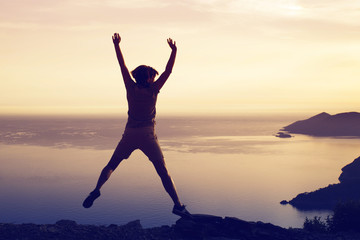 Fototapeta premium Silhouette young woman hovered in a jump in the air against the seascape and mountains at sunset, rear view, toned. Concept of freedom, happiness, success.