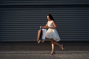 Shopping. Beautiful fashionable woman wearing white stylish dress curiously looking into shopping bags. Grey wall on background.