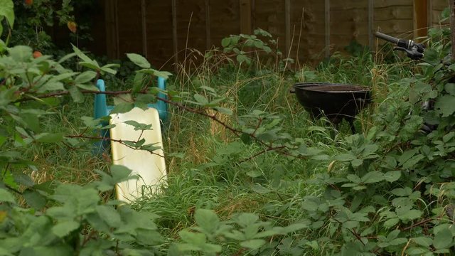 Overgrown garden with plastic slide and rusty BBQ as bramble moves in the wind
