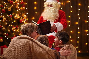 Santa Claus reading book for family. Mother and children sitting indoor near decorated xmas tree with lights - Merry Christmas and Happy Holidays!