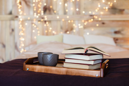 Stack of books and two cups of tea on a bed with background of lights. Brown bedroom with wooden wall. Romantic evening with books