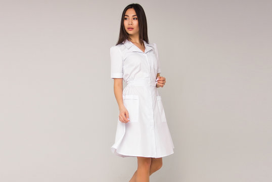 young stylish brunette nurse with long hair in white medical dress is standing and looking away on the white wall background. medical fashion concept. free space
