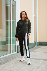 young glamour brunette girl in black sports costume is standing casual  with mobile phone in hand and looking straight near windows background outdoor, lifestyle concept, free space, place for text