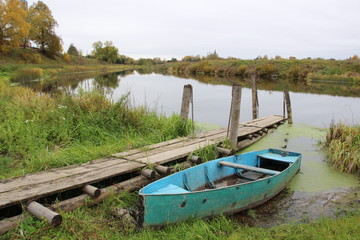 Naklejka na ściany i meble Blue fishing boat hang to old wooden gangway with background of autumn river landscape