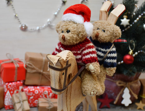 Two Teddy Bears are riding a wooden hobbyhorse on a background of a Christmas tree & boxes with Christmas presents