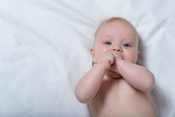 Portrait of a cute blond baby with a hand in his mouth. White sheet. Top view