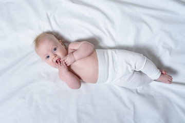 Cute blond baby with fingers in his mouth lies on a white sheet. Top view