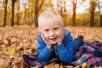 Cute little blond boy lies on a plaid, yellow autumn leaves. Smiling and having fun. Fall day