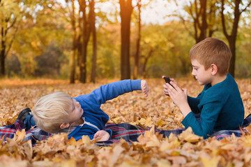 Two little brothers take pictures of each other, lying in yellow autumn leaves. Fall day