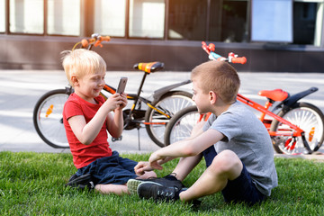 Two boys are photographed on a smartphone while sitting on the grass. Rest after cycling, bicycles in the background