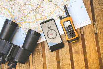 Still life view from above of adventurous objects. Walkie talkie, binoculars and electronic compass on smartphone over a map, ready for planning a new travel route. Travelling, tech adventure concept