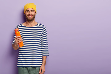 Studio shot of handsome smiling hipster guy with dark bristle, wears yellow hat and casual striped jumper, holds flask of hot drink, enjoys spare time, isolated over purple wall, free space on right