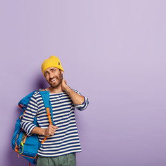 Vertical image of fatigue European man touches neck, suffers from pain in neck, dressed in casual wear, carries backpack on shoulder, smirks face, shows white teeth, isolated over purple background