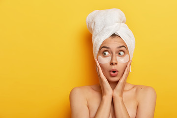 Photo of surprised young Afro woman touches cheeks with hands, looks aside with stupor, wears wrapped towel, applies white patches under eyes, models over yellow background, has spa procedure